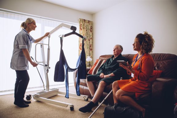 A caregiver assists a man in a living room while a woman sits nearby.